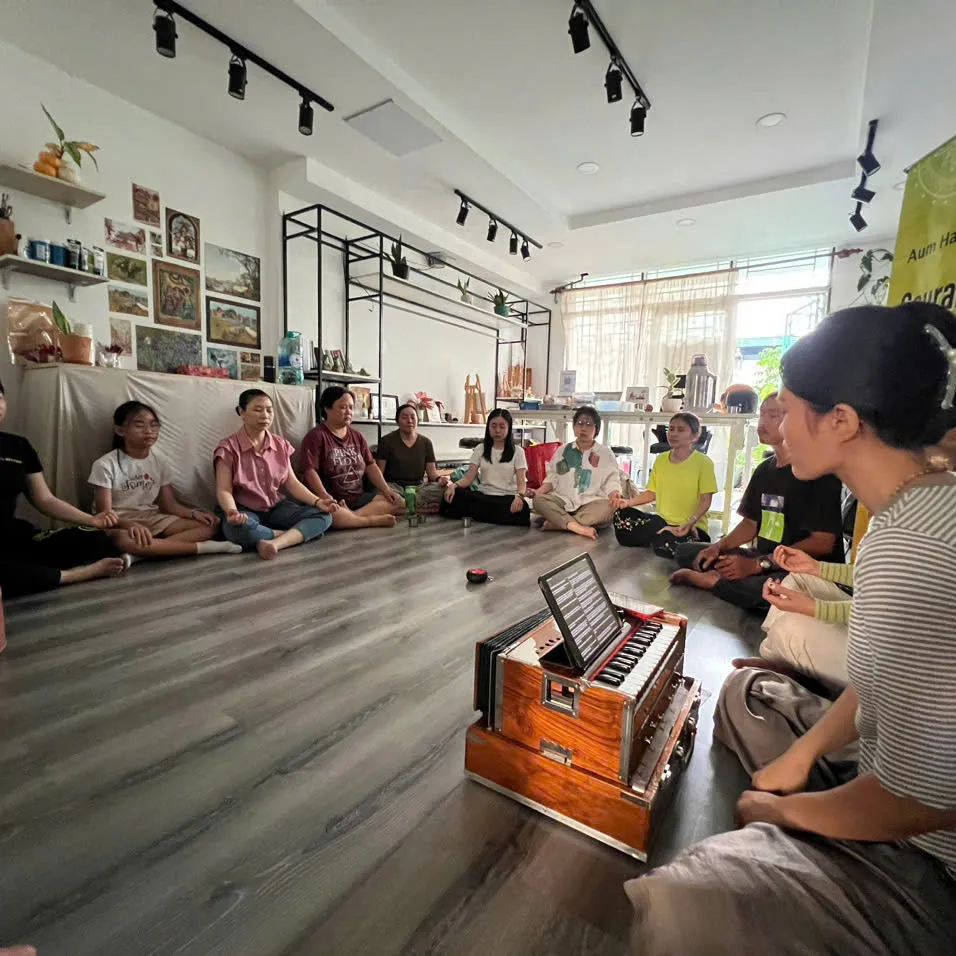 A circle of people in a studio, one playing the harmonium.