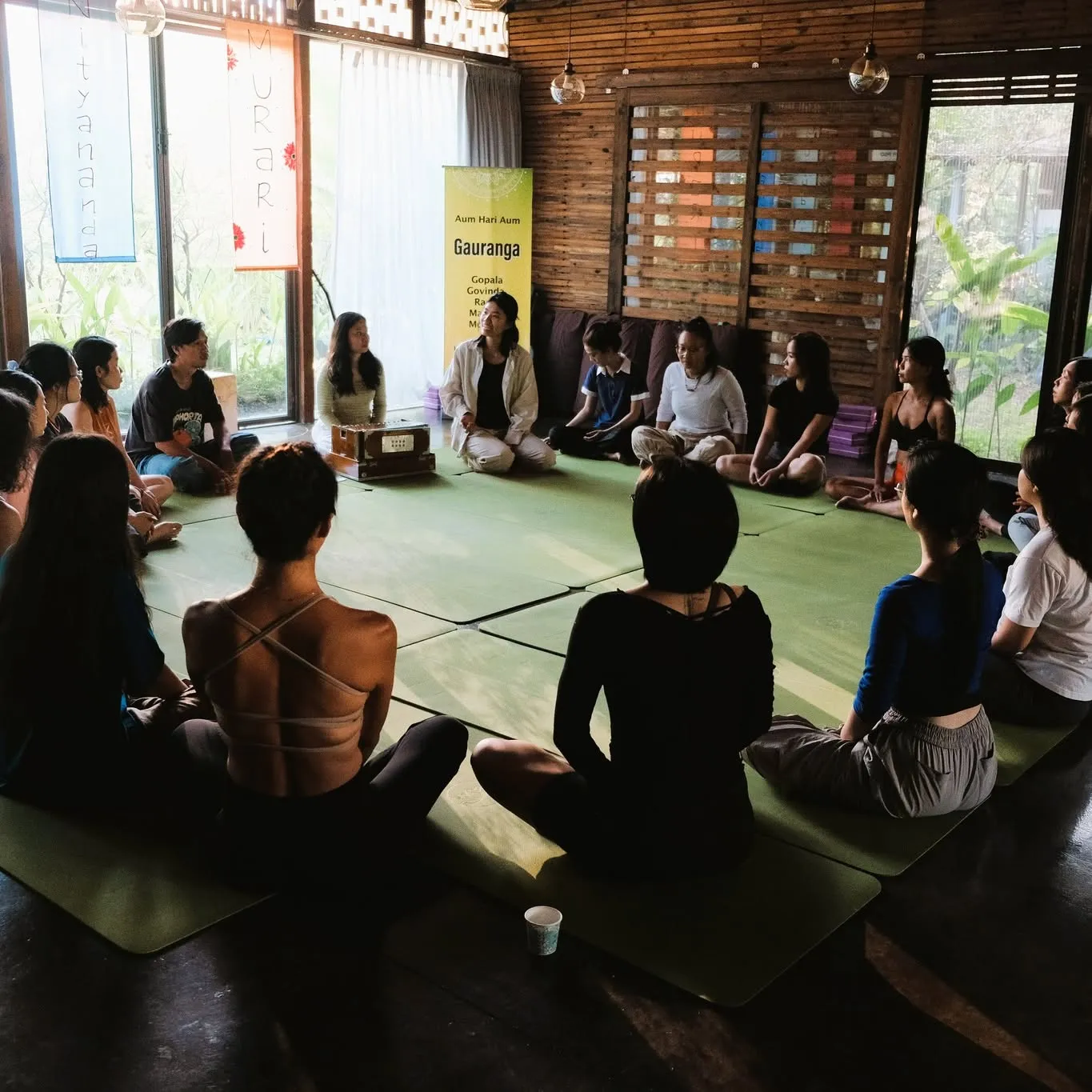 Một vòng tròn người ngồi trong nhà gỗ sáng sủa, có người chơi nhạc ở giữa.
