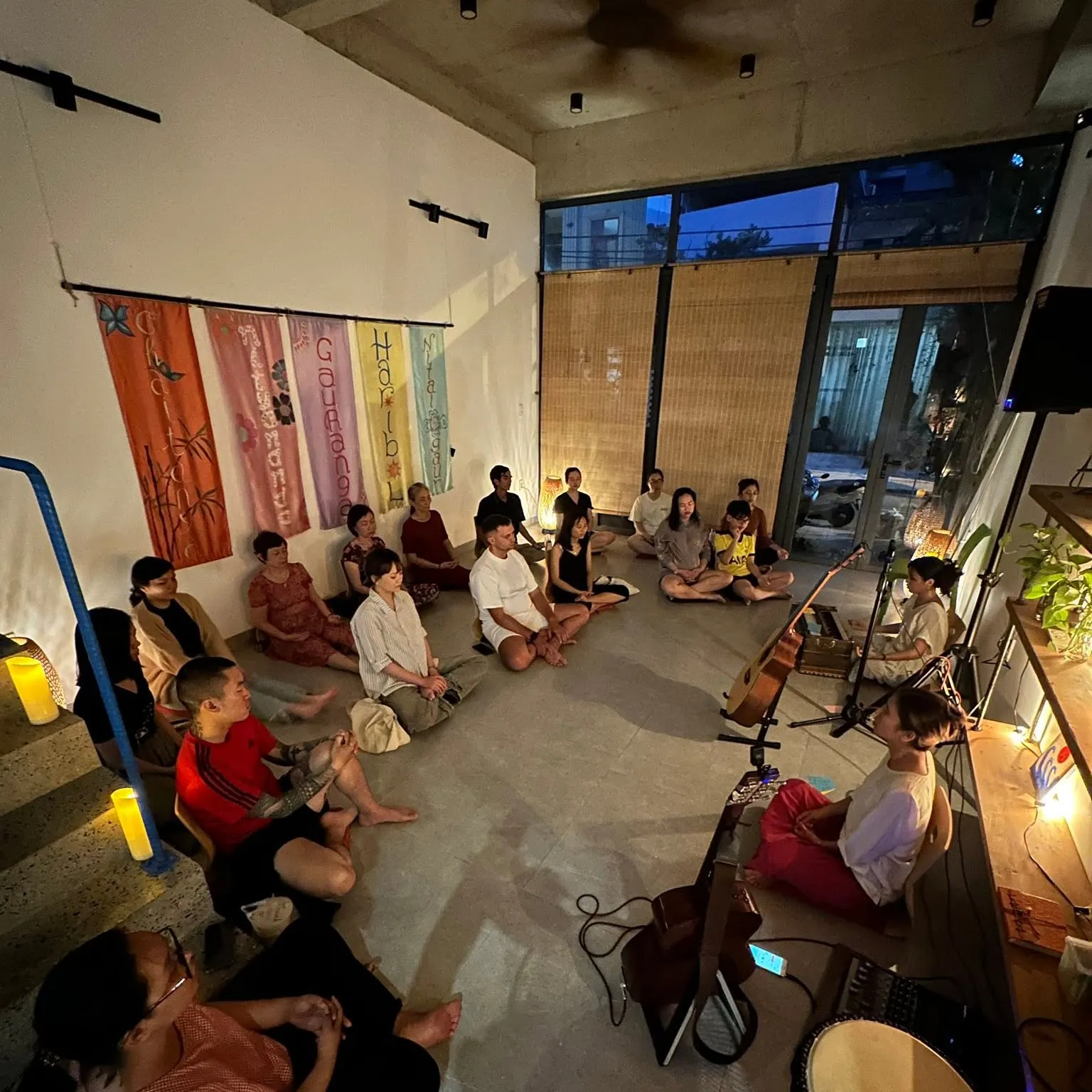Một vòng tròn đông người ngồi trên sàn, cùng hát dưới ánh đèn ấm.