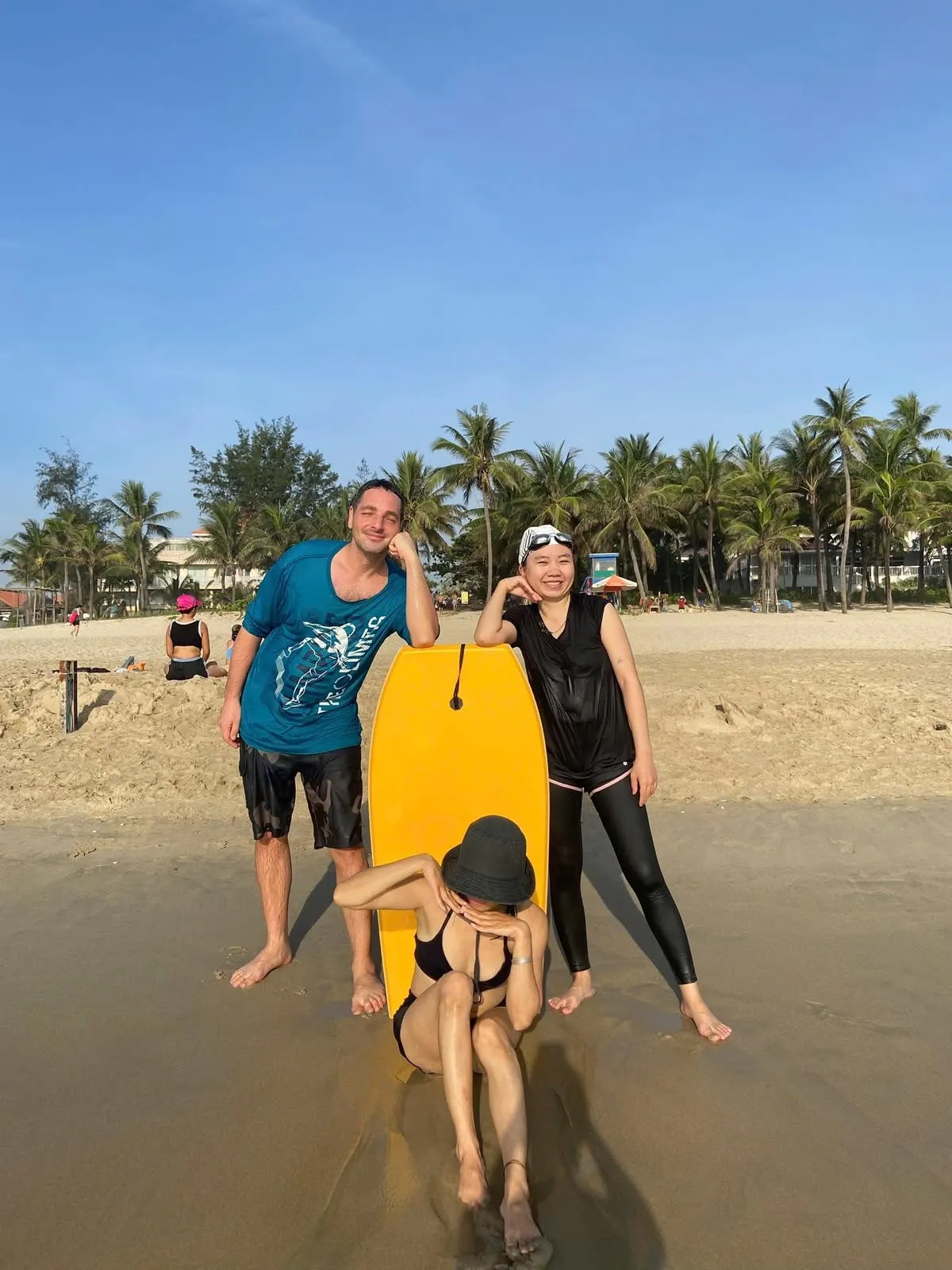 Ba người cười đùa tạo dáng bên một tấm ván lướt sóng màu vàng trên bãi biển.