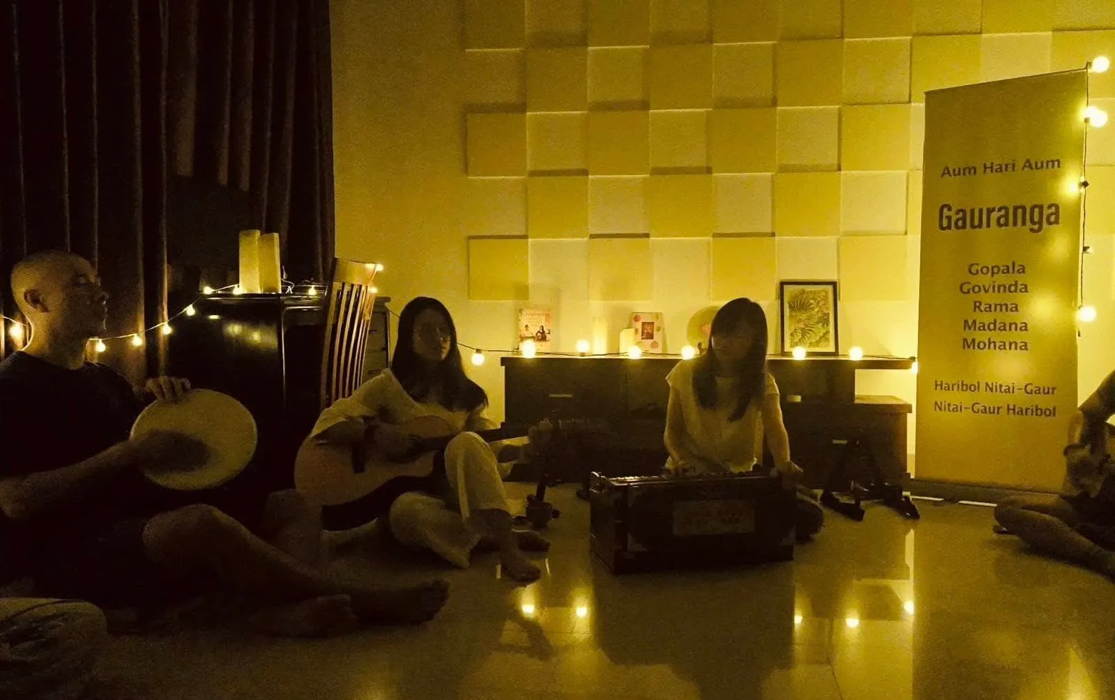 Ba người chơi trống, guitar và đàn harmonium dưới ánh đèn ấm, bên tấm băng rôn thần chú.