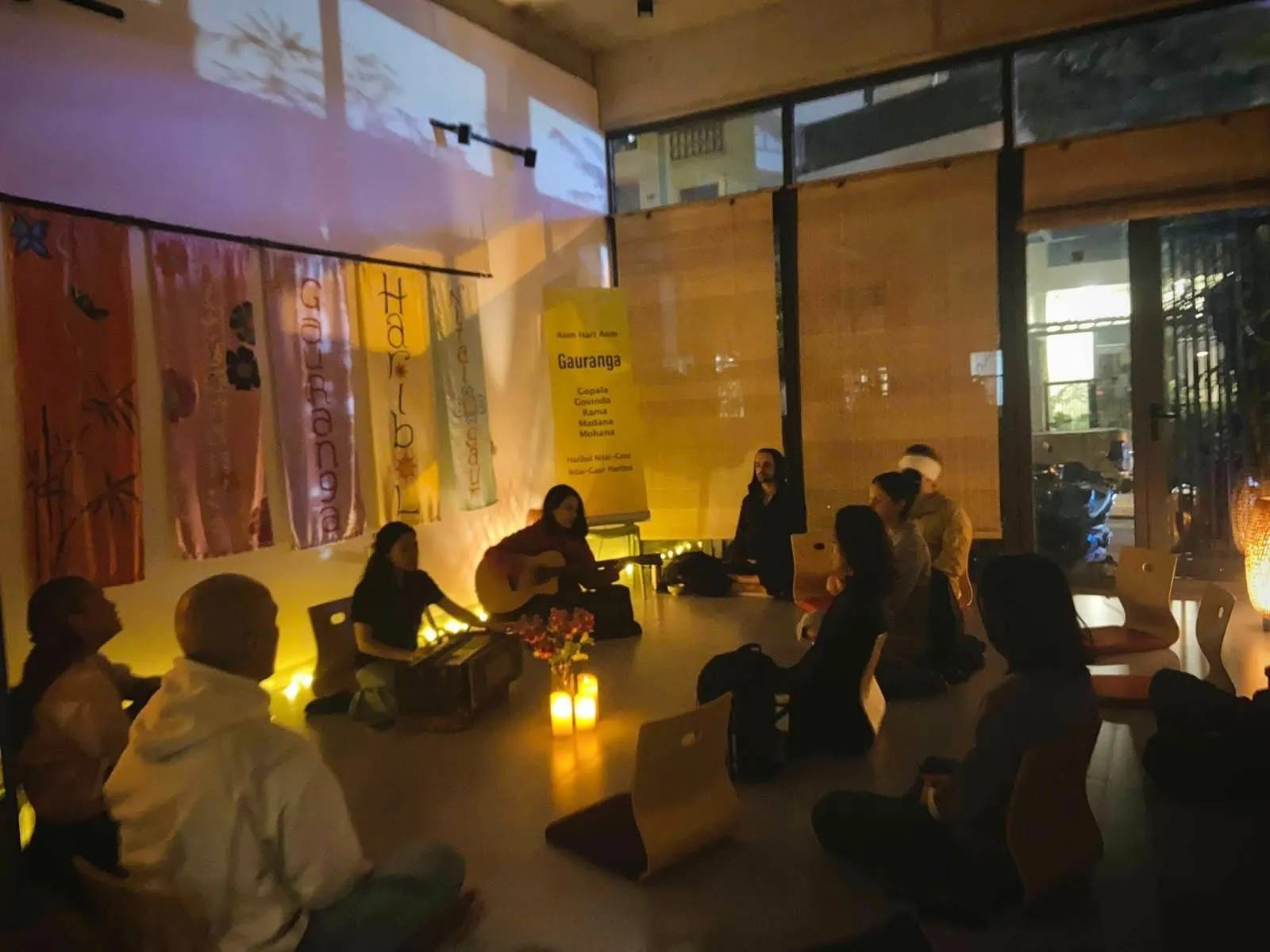 Một vòng tròn người ngồi thiền trong căn phòng ánh nến, bên đàn harmonium, cây đàn guitar và những tấm vải thần chú.